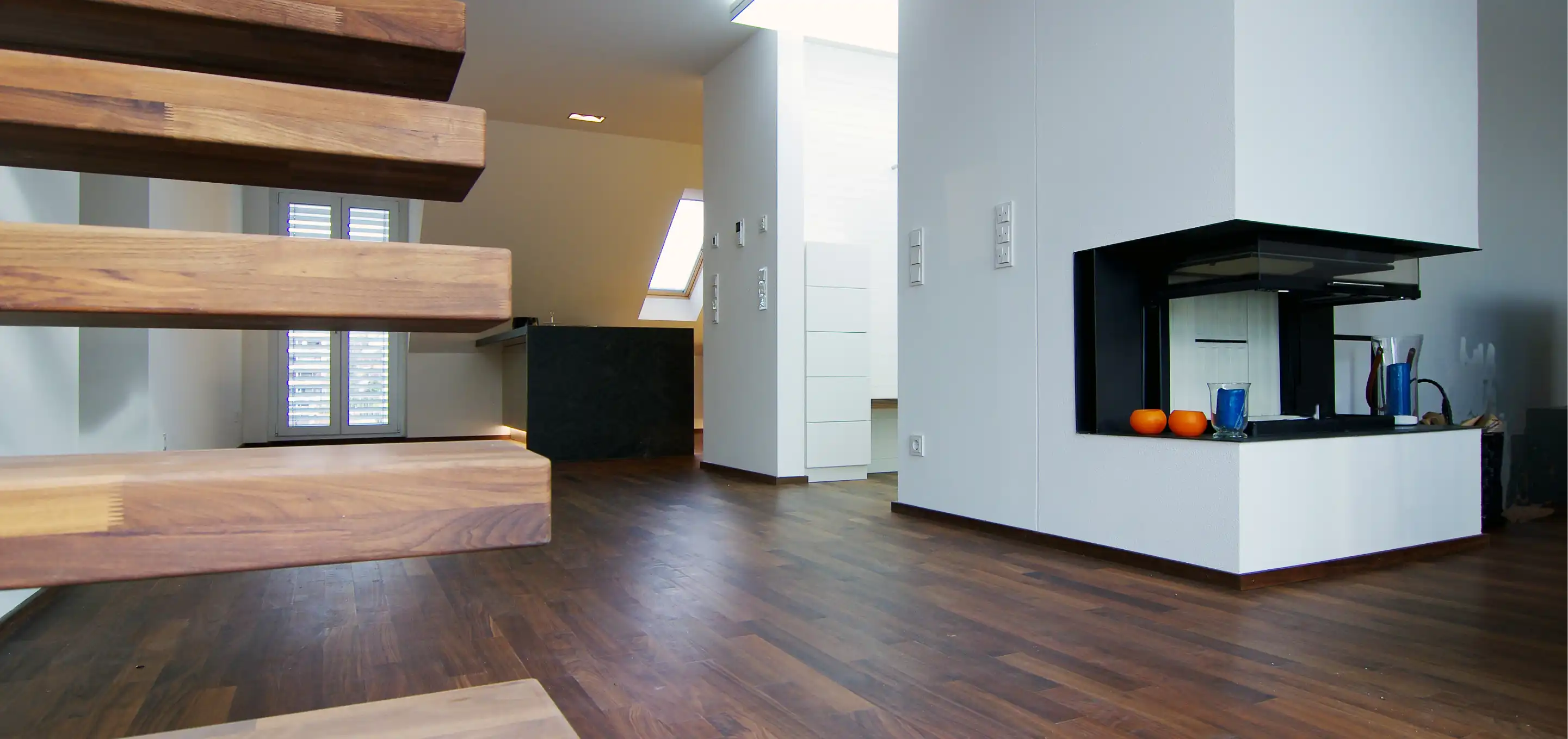 Wohnbereich unter der Dachschräge mit Kamin, Holztreppe ohne Geländer und dunklen Holzdielen